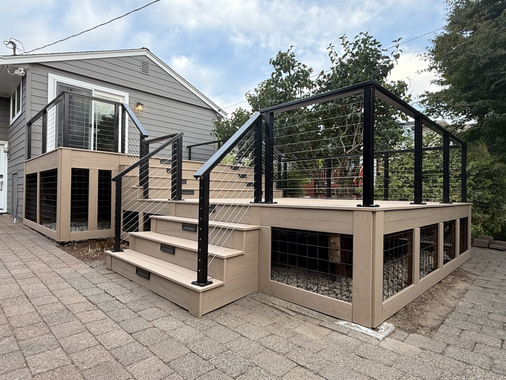 Olympic Decks Multi-Level Beige Composite Deck With Black Cable Railings And Under-Deck Wire Mesh Skirting Next To A Paver Patio.